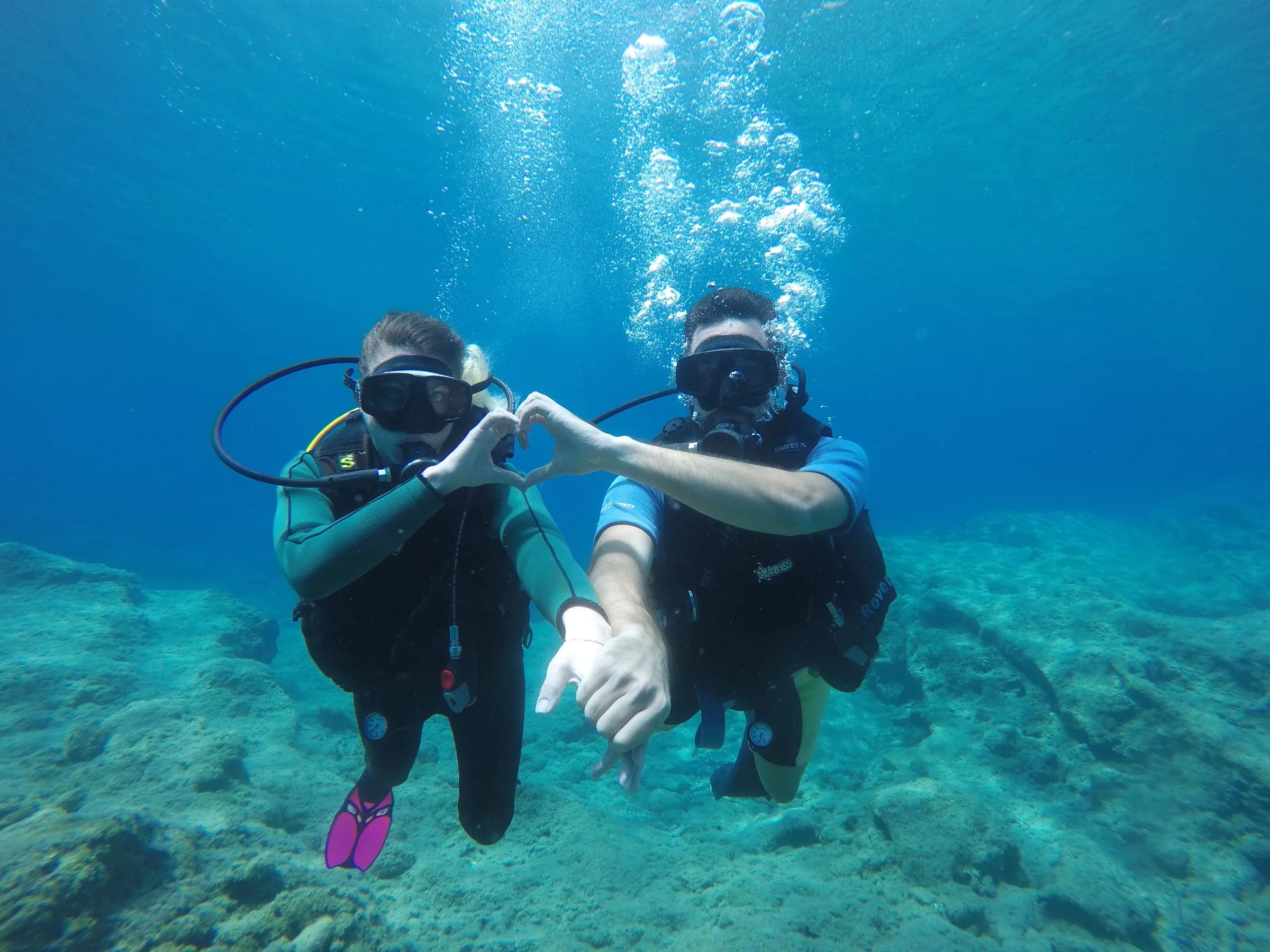 Two scuba divers making a heart shape with their hands above a rocky seabed in clear blue water