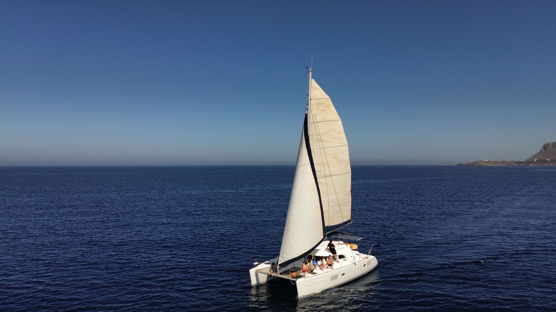 Sailing catamaran with white sails on a calm blue sea, passengers relaxing on deck and a rocky coast in the distance