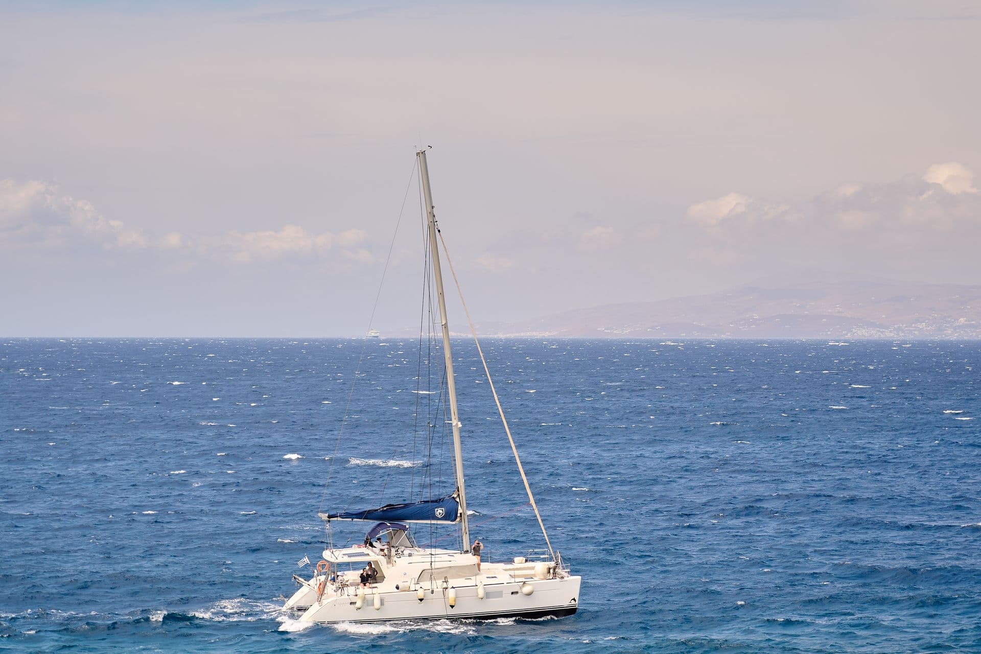 White sailing catamaran with its mainsail furled crossing a choppy blue sea, a hilly coastline on the horizon