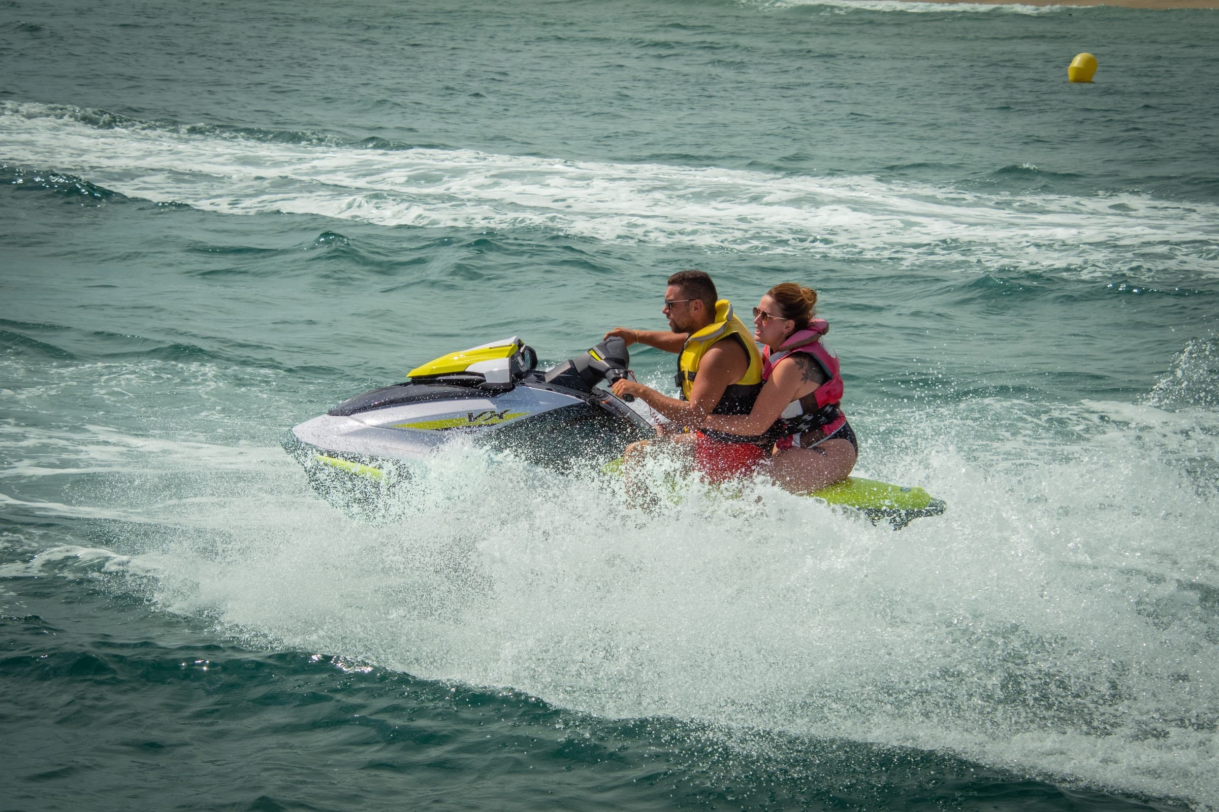 Man and woman in life jackets riding a white and yellow jet ski through spray, a yellow buoy in the background