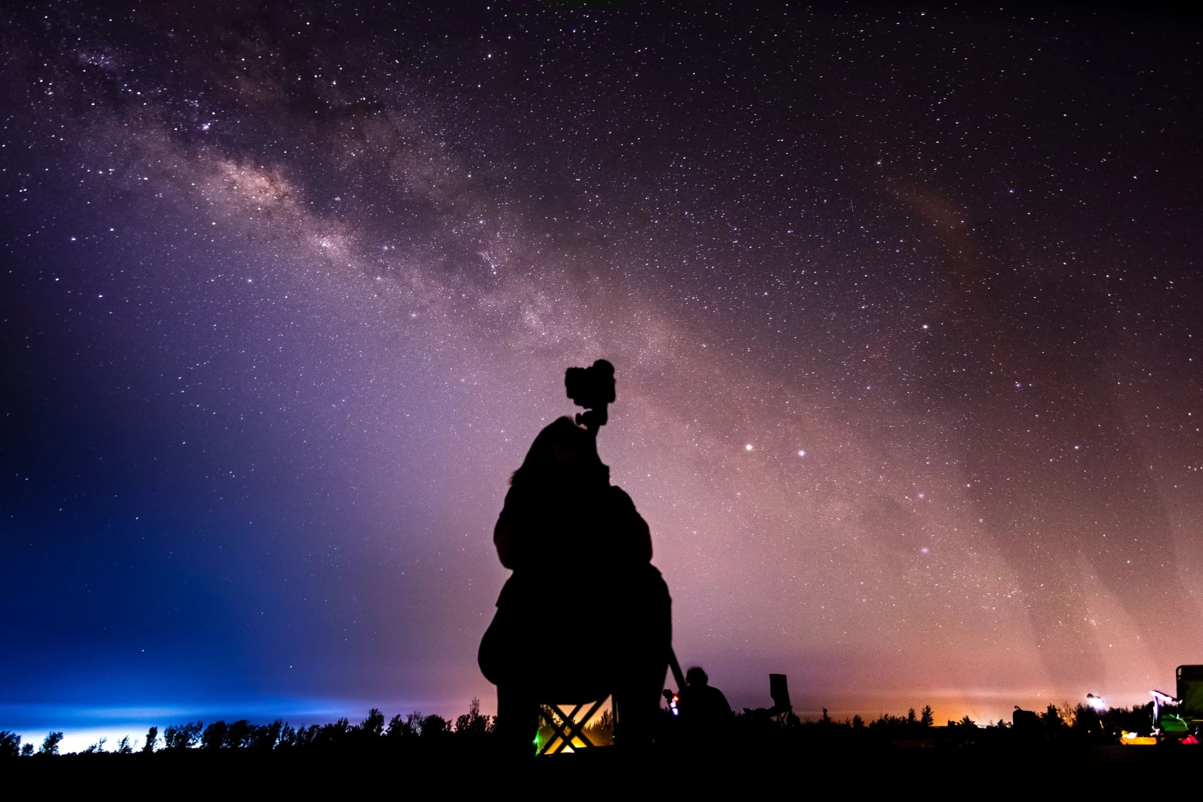 Silhouette of a photographer with a camera on a tripod under the Milky Way, with other stargazers and folding chairs nearby