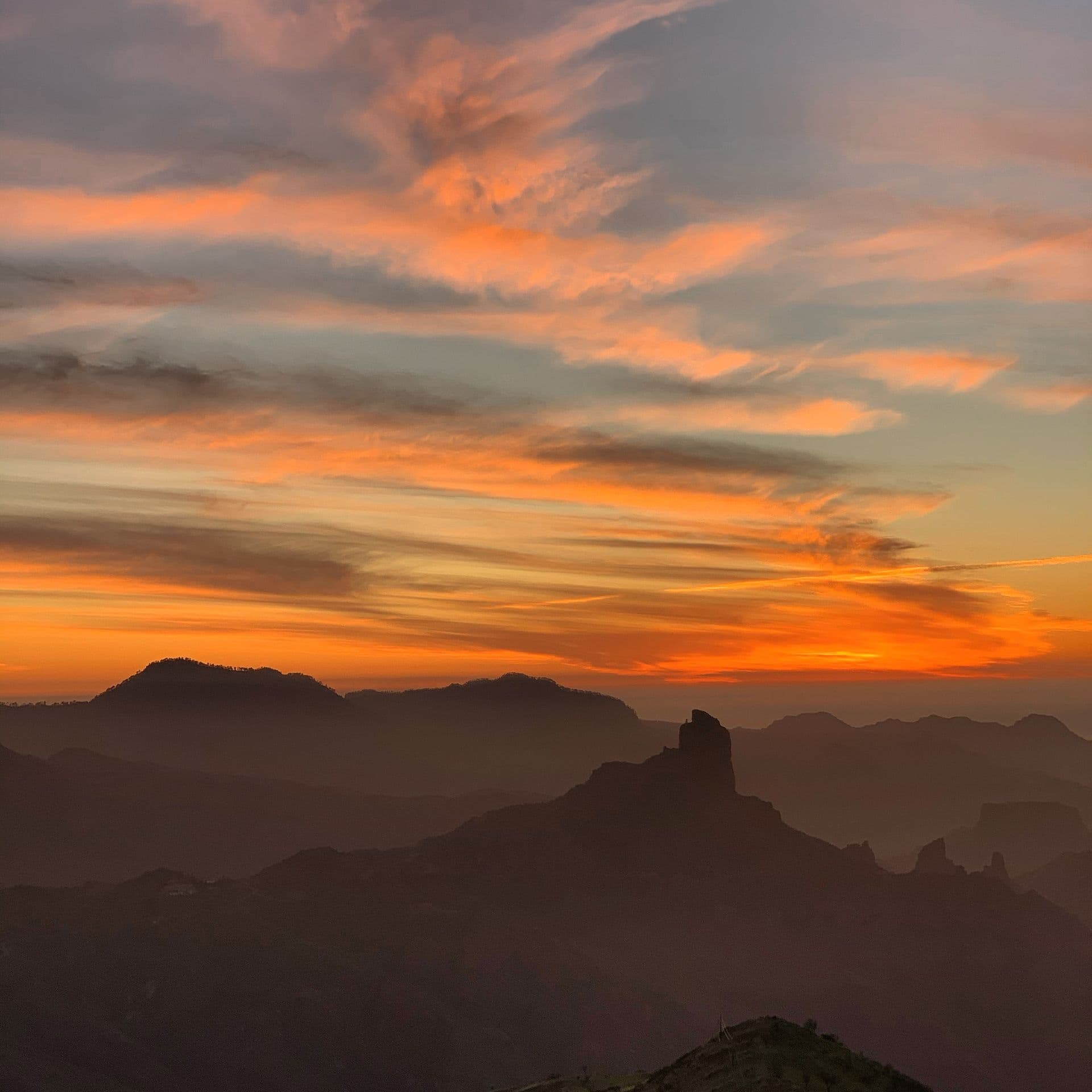 Tall rock pillar and layered mountain ridges silhouetted against an orange sunset sky with streaked clouds