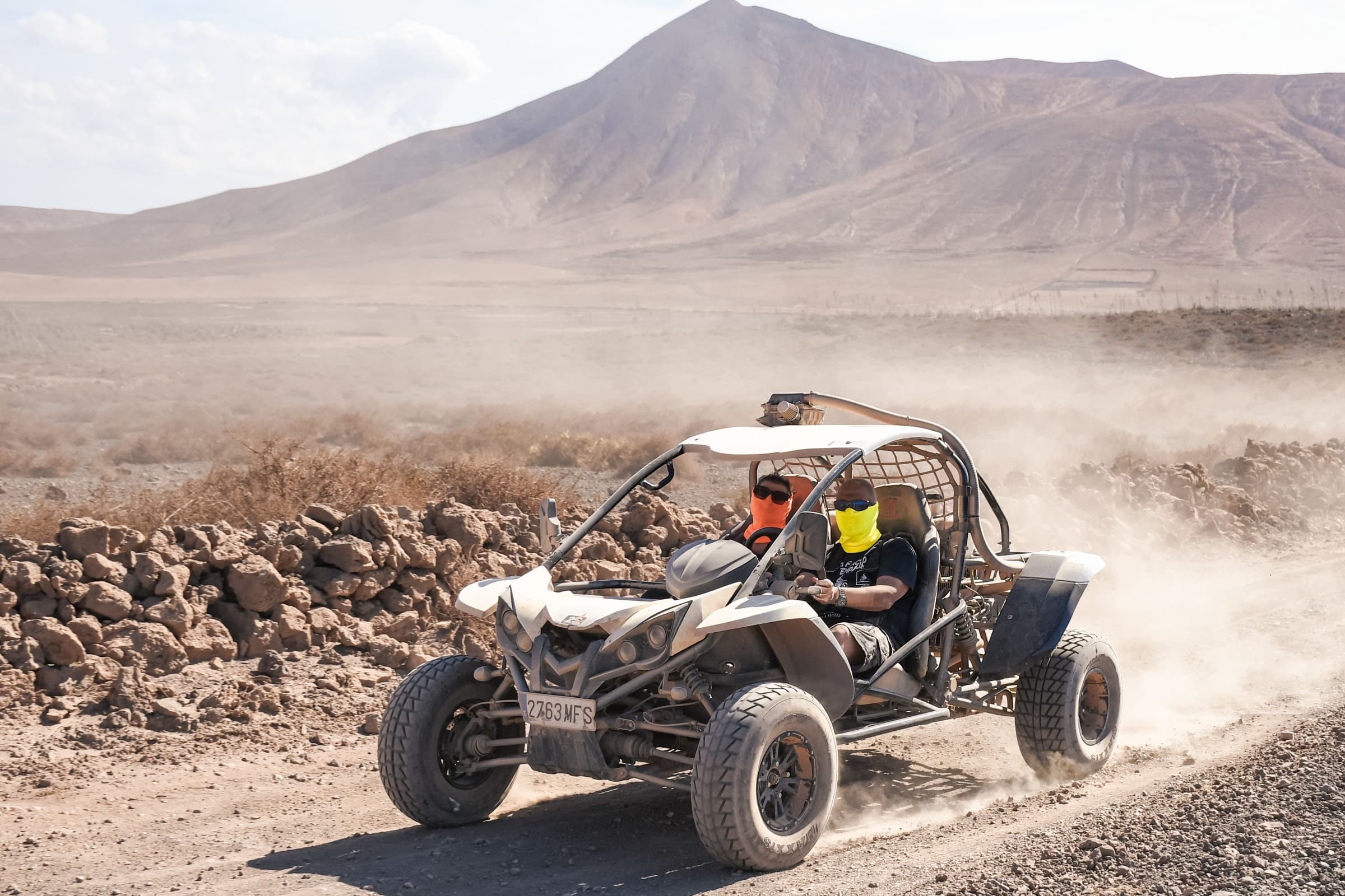 Off-road buggy with two riders in face buffs and sunglasses raising dust on a dirt track below a bare volcanic mountain
